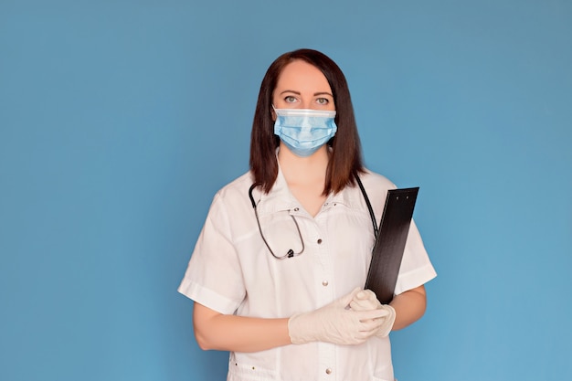 Female doctor in medical mask with a stethoscope writes on a tablet on a blue background, copy space