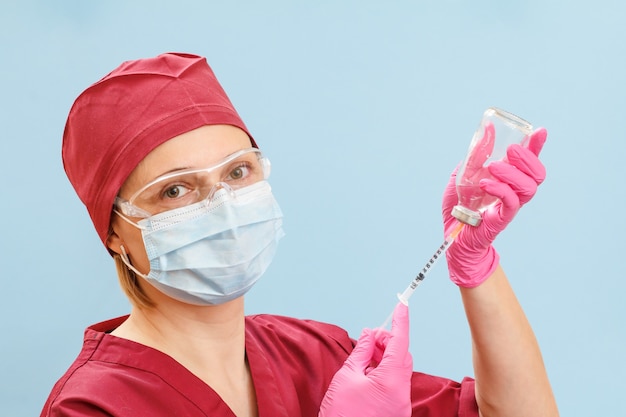 Female doctor in a medical uniform, a ptotective mask and latex gloves holding a glass bottle and filling in a syringe with liquid medicine for injection on the blue surface