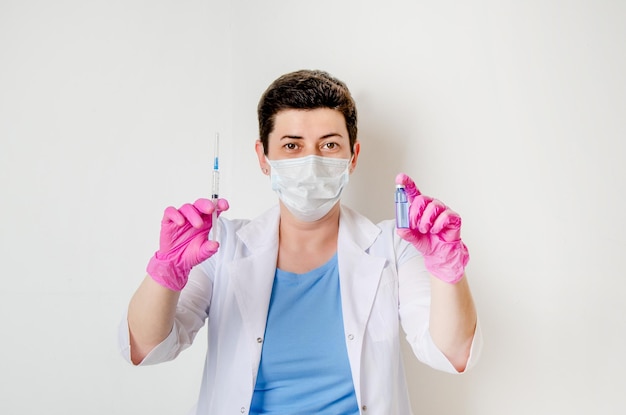 Female doctor in protective mask gloves and medical gown with syringe and an injection bottle