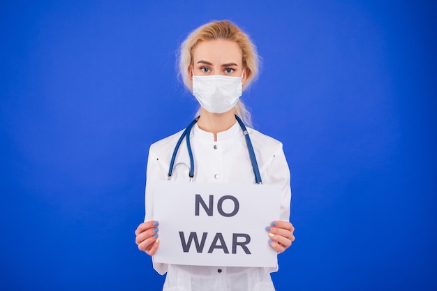 A female doctor in a protective mask holds a no war poster on a blue background