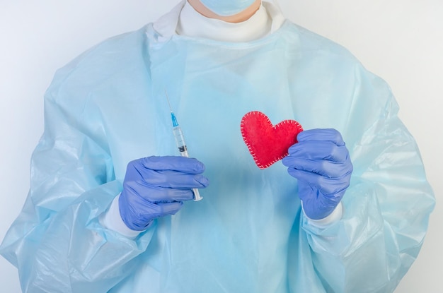 A female doctor in a protective suit holds a red heart and a syringe in her hands on a white background