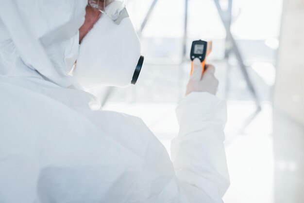 Female doctor scientist in lab coat defensive eyewear and mask standing indoors with infrated thermometer