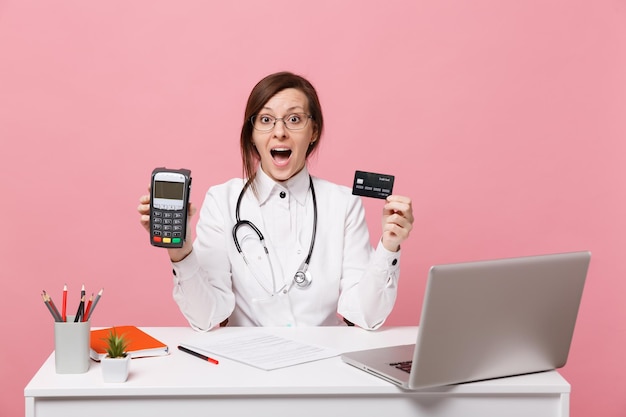 Female doctor sit at desk work on computer with medical document credit card in hospital isolated on pastel pink wall background. Woman in medical gown glasses stethoscope. Healthcare medicine concept