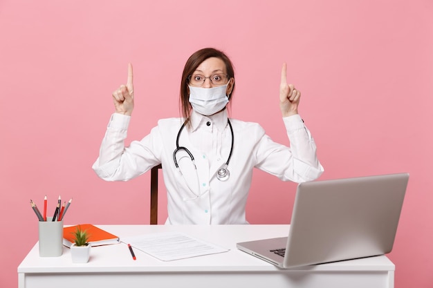 Female doctor sit at desk work on computer with medical document in face mask in hospital isolated on pastel pink wall background. Woman in medical gown glasses stethoscope Healthcare medicine concept
