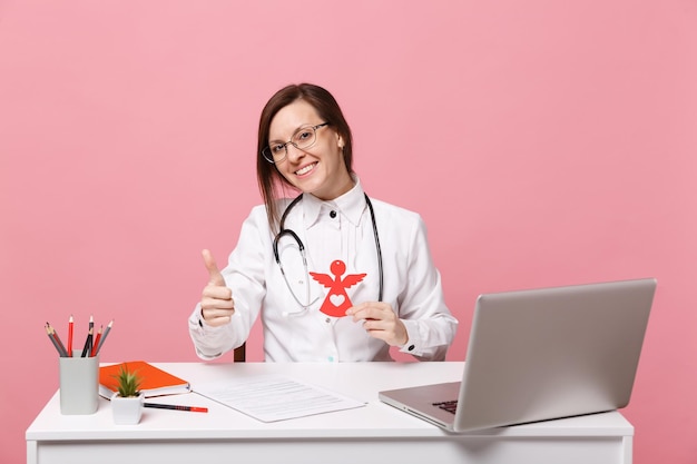 Female doctor sit at desk work on computer with medical document hold angel in hospital isolated on pastel pink wall background. Woman in medical gown glasses stethoscope. Healthcare medicine concept.