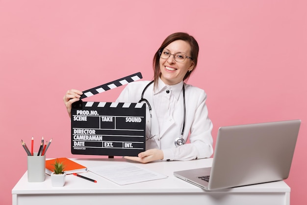 Female doctor sit at desk work on computer with medical document hold board in hospital isolated on pastel pink wall background. Woman in medical gown glasses stethoscope. Healthcare medicine concept.