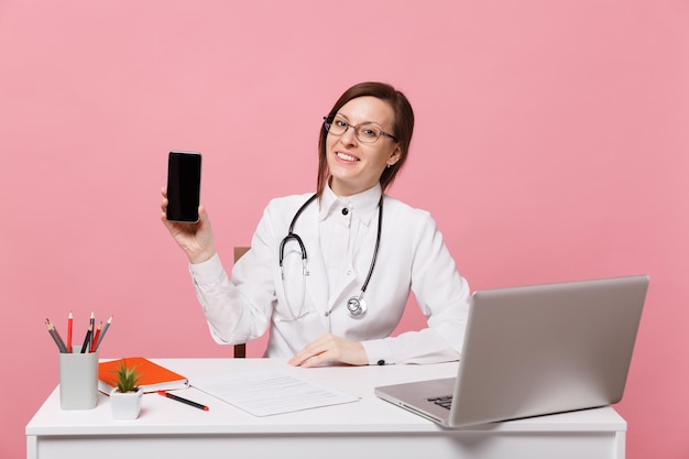 Female doctor sit at desk work on computer with medical document hold cellphone in hospital isolated on pastel pink background. Woman in medical gown glasses stethoscope. Healthcare medicine concept.