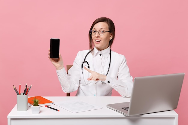 Female doctor sit at desk work on computer with medical document hold cellphone in hospital isolated on pastel pink background. Woman in medical gown glasses stethoscope. Healthcare medicine concept.
