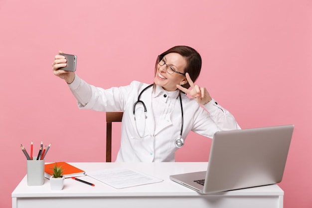 Female doctor sit at desk work on computer with medical document hold cellphone in hospital isolated on pastel pink background. Woman in medical gown glasses stethoscope. Healthcare medicine concept.