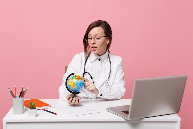 Female doctor sit at desk work on computer with medical document hold globe in hospital isolated on pastel pink wall background. Woman in medical gown glasses stethoscope. Healthcare medicine concept.