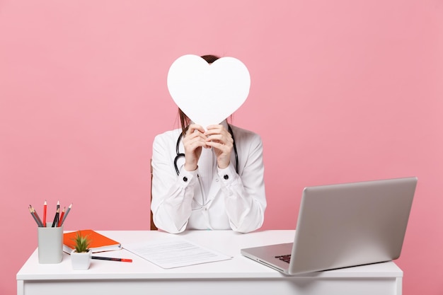 Female doctor sit at desk work on computer with medical document hold heart in hospital isolated on pastel pink wall background. Woman in medical gown glasses stethoscope. Healthcare medicine concept.