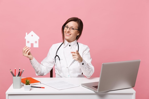 Female doctor sit at desk work on computer with medical document hold house in hospital isolated on pastel pink wall background. Woman in medical gown glasses stethoscope. Healthcare medicine concept.