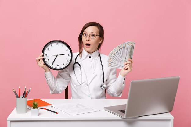 Female doctor sit at desk work on computer with medical document hold money in hospital isolated on pastel pink wall background. Woman in medical gown glasses stethoscope. Healthcare medicine concept.