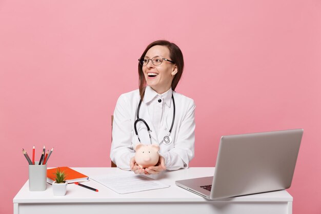 Female doctor sit at desk work on computer with medical document hold pig money in hospital isolated on pastel pink background. Woman in medical gown glasses stethoscope. Healthcare medicine concept.