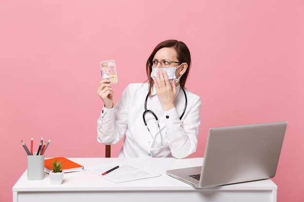 Female doctor sit at desk work on computer with medical document hold pills in hospital isolated on pastel pink wall background. Woman in medical gown glasses stethoscope. Healthcare medicine concept.