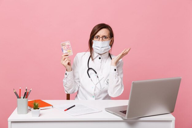 Female doctor sit at desk work on computer with medical document hold pills in hospital isolated on pastel pink wall background. Woman in medical gown glasses stethoscope. Healthcare medicine concept.