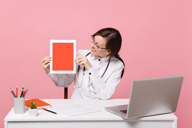 Female doctor sit at desk work on computer with medical document hold tablet in hospital isolated on pastel pink wall background. Woman in medical gown glasses stethoscope. Healthcare medicine concept