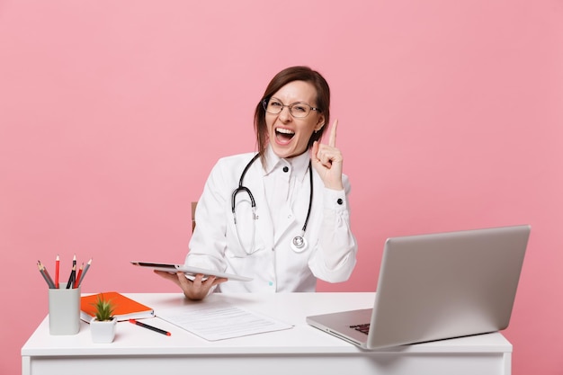 Female doctor sit at desk work on computer with medical document hold tablet in hospital isolated on pastel pink wall background. Woman in medical gown glasses stethoscope. Healthcare medicine concept