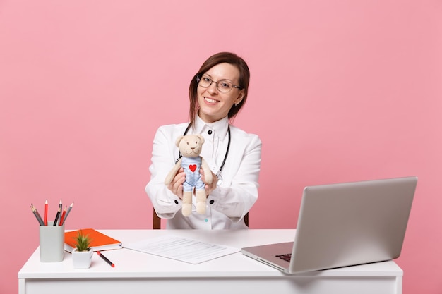 Female doctor sit at desk work on computer with medical document hold toy in hospital isolated on pastel pink wall background. Woman in medical gown glasses stethoscope. Healthcare medicine concept.