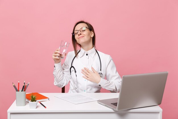 Female doctor sit at desk work on computer with medical document hold water in hospital isolated on pastel pink wall background. Woman in medical gown glasses stethoscope. Healthcare medicine concept.