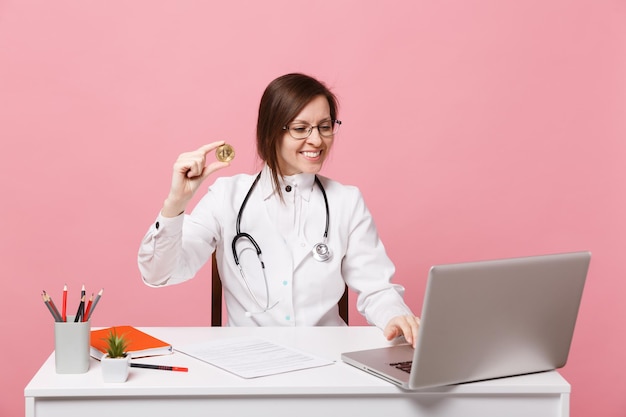 Female doctor sits at desk works on computer with medical document hold coin in hospital isolated on pastel pink wall background. Woman in medical gown glasses stethoscope. Healthcare medicine concept