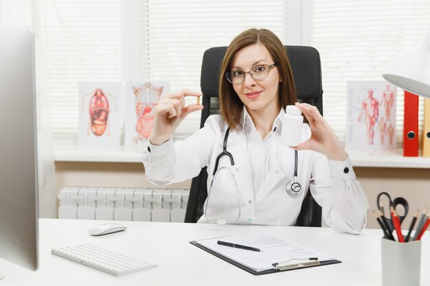 Female doctor sitting at desk, holding bottle with white pills, working with medical documents in light office in hospital