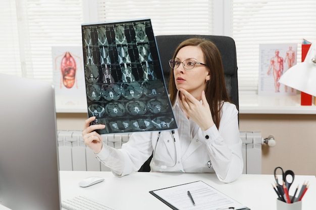 Female doctor sitting at desk with computer, film x-ray the brain by radiographic image ct scan mri in light office in hospital