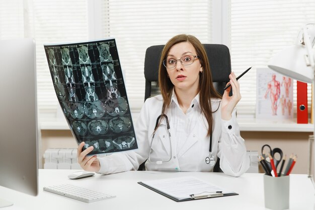 Female doctor sitting at desk with computer, film x-ray the brain by radiographic image ct scan mri in light office in hospital