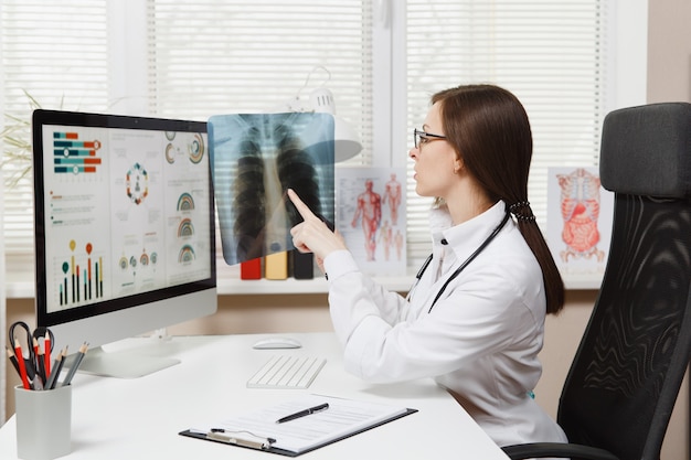 Female doctor sitting at desk, working on computer, holding X-ray of lungs, fluorography, roentgen in light office in hospital