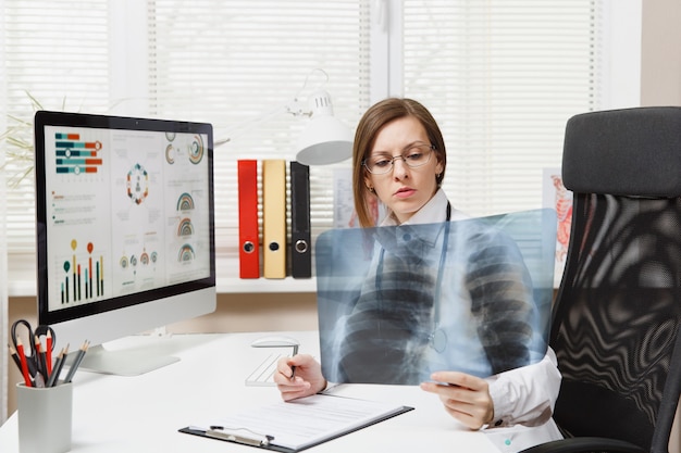 Female doctor sitting at desk, working on computer, holding X-ray of lungs, fluorography, roentgen in light office in hospital