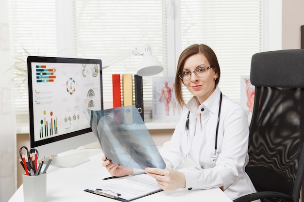 Female doctor sitting at desk, working on computer, holding X-ray of lungs, fluorography, roentgen in light office in hospital