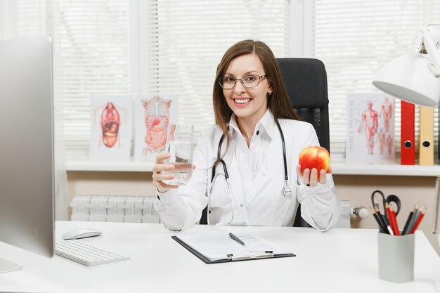 Female doctor sitting at desk, working on computer with medical documents in light office in hospital