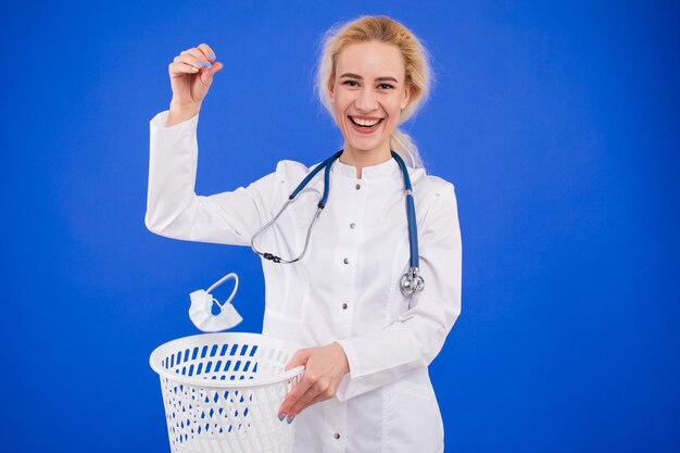 Женщина-врач выбрасывает защитную маску в мусорное ведро на синем фоне