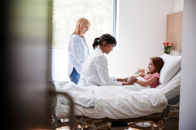 Female Doctor Visiting Mother And Daughter Lying In Bed In Hospital Ward