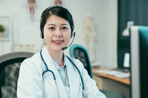 Female doctor wearing headset while using computer at desk in\
clinic office. beautiful woman medical staff consult client patient\
on microphone and headphone. lady hospital worker face camera\
smiling