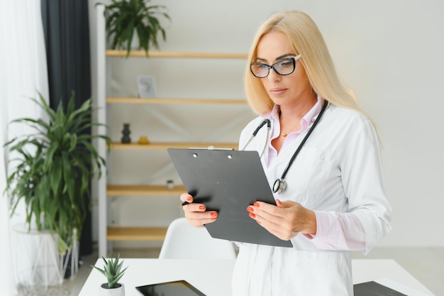 Female doctor wearing lab coat and stethoscope and holding clipboard in her hands while standing at hospital