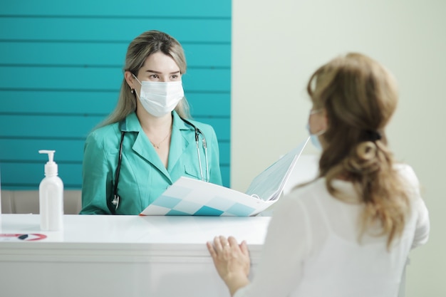 병원의 리셉션 영역에서 여성 환자와 이야기하는 의료 마스크를 쓰고 여성 의사
