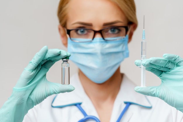 Female doctor wearing protective mask and gloves and holding bottle of vaccine medicine and syringe