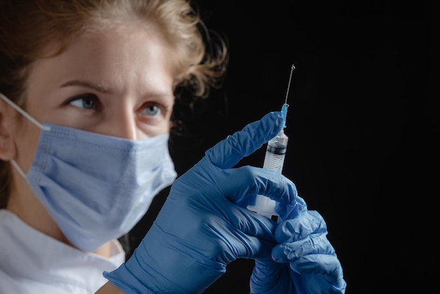 A female doctor wearing a protective mask and gloves holds a syringe with a vaccine or injection aga