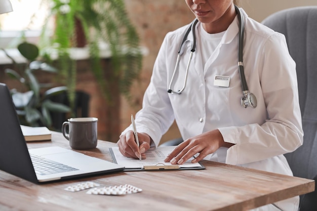 Female doctor wearing white coat sitting at desk in loft office doing paperwork making notes in medi