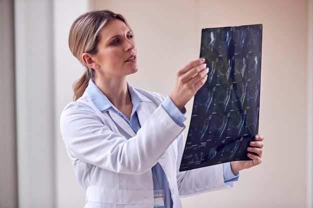 Female Doctor Wearing White Coat Standing In Hospital Corridor Looking At CT Or MRI Scan