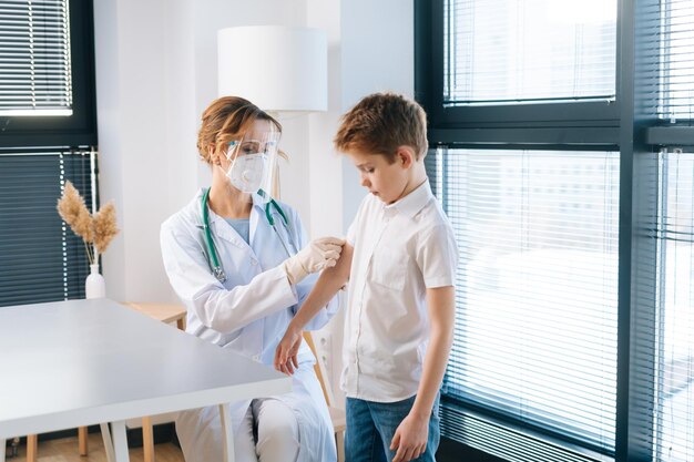 Female doctor wearing white uniform and safety mask applying plaster on shoulder of child boy after vaccination injection by window