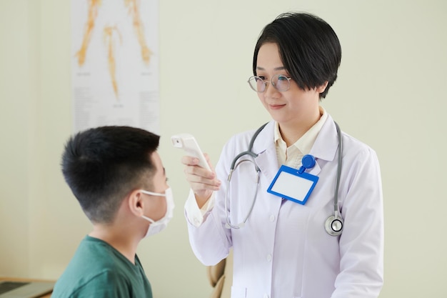 Female doctor in white coat measuring the temperature of little boy with medical equipment during medical exam at hospital