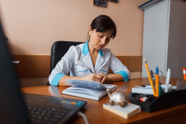 A female doctor in white medical uniform is sitting at a wooden table in the office and makes up a work schedule for the staff. Working days. The doctor works