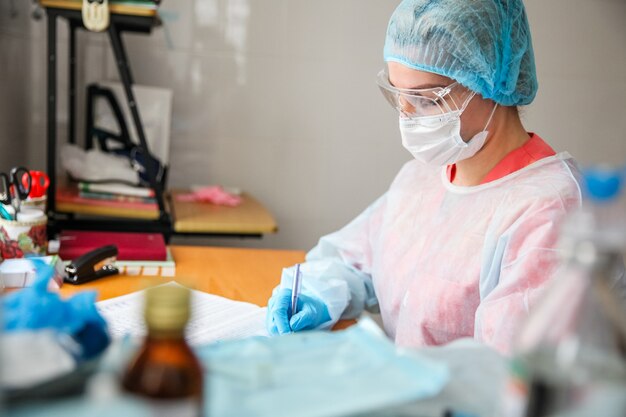 A female doctor in a white protective suit, blue cap, blue gloves and mask sits at a table and fills out a magazine