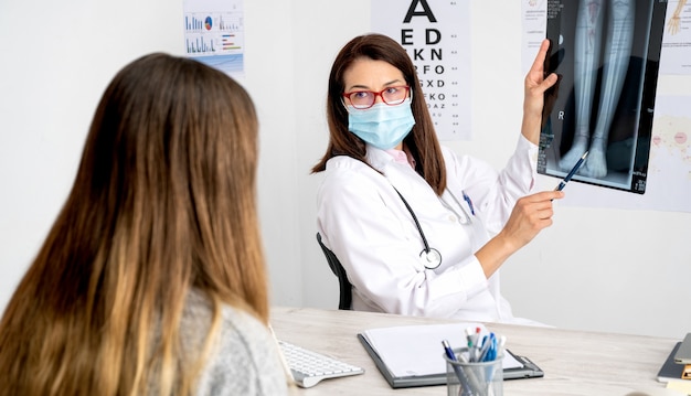 X線を示す彼女の診察で患者に付き添うマスクを持つ女性医師