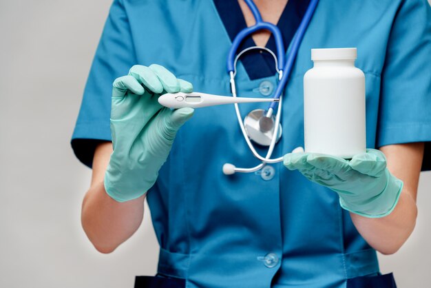 Female doctor with stethoscope wearing protective mask and latex gloves over light grey background holding thermometer and can jar of medicine pills