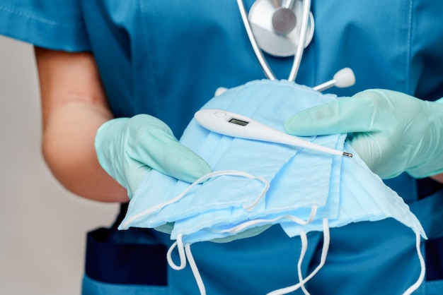 Female doctor with stethoscope wearing protective mask and latex gloves over light grey background holding thermometer