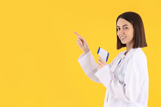 Female doctor with thermometer gun on yellow background