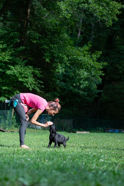 Female dog trainer luring young black labrador retriever puppy with food to teach her basic obedience outside in green nature